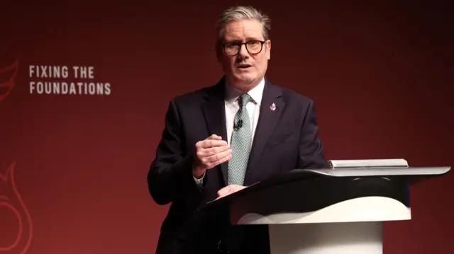 Sir Keir Starmer stands at a podium in front of a dark red background with the words "Fixing the foundations" written on it. Starmer is wearing a black suit with a turquoise and white tie. He has a red remembrance poppy pinned to his suit