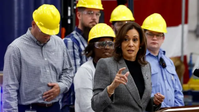 Kamala Harris speaking at a stand. Behind her stand four men and a woman wearing yellow hard hats