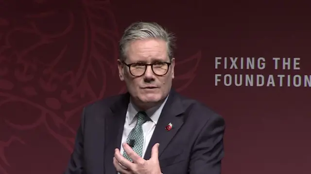 Mid-shot of Starmer as he answers questions. He's leaning on podium, his left hand raised with fingers apart in front of him. He's wearing dark suit, white shirt and patterned tied