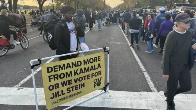 A man with a sign saying 'demand more from Kamala'