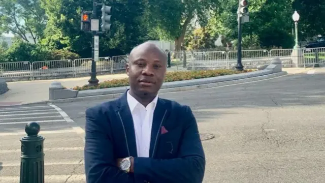 Dr Yves Kaduli stands near street crossing. He's wearing a dark suit jacket with metal details on lapels and handkerchief in pocket.