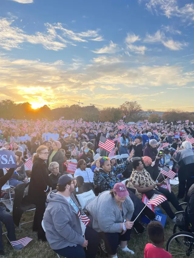 Crowd of people with american flags sit in the twilight , very packed, on a lawn