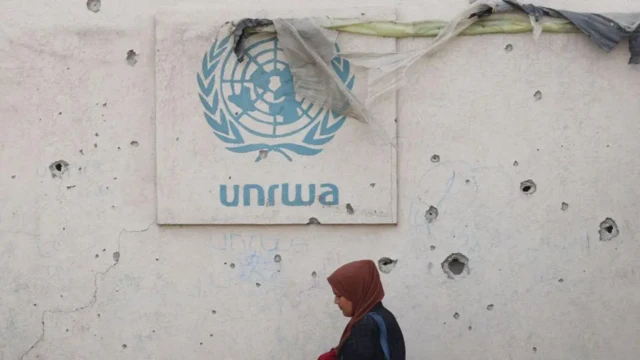 A woman in a hijab walks past a concrete wall riddled with bullet holes bearing the blue unwra logo