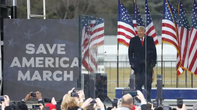 Trump standing on a stage, talking to supporters, with a screen showing the text 'SAVE AMERICA MARCH'