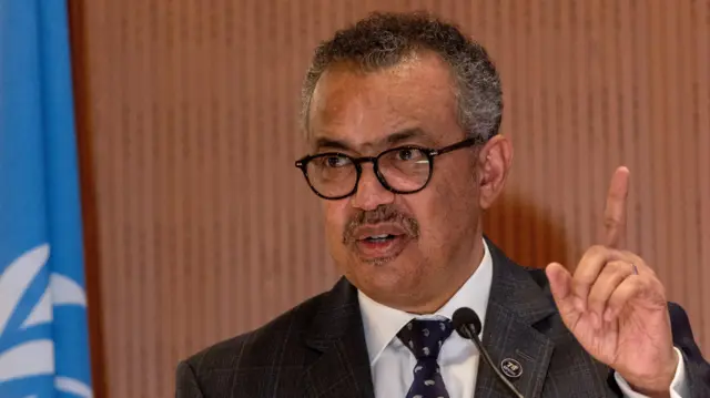 Dr. Tedros Adhanom Ghebreyesus stands in a dark grey suit and tie, he is wearing glasses speaking into a podium microphone with his hand pointed upwards