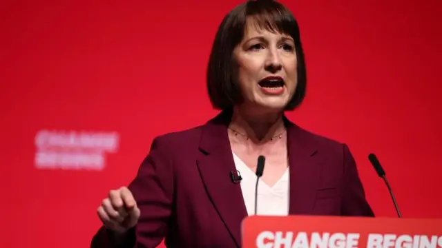 Reeves points as she delivers her address in front of a red backdrop