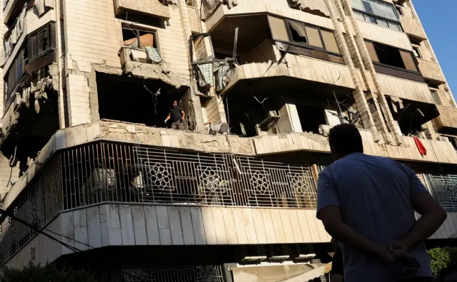 A man with his hands behind his back and his back to the camera looks up at an apartment block which has been hit by an airstrike