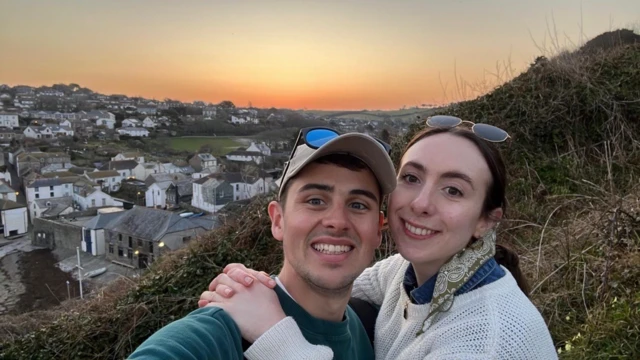 Ross and his girlfriend pose on a hillside in front of a sunset.