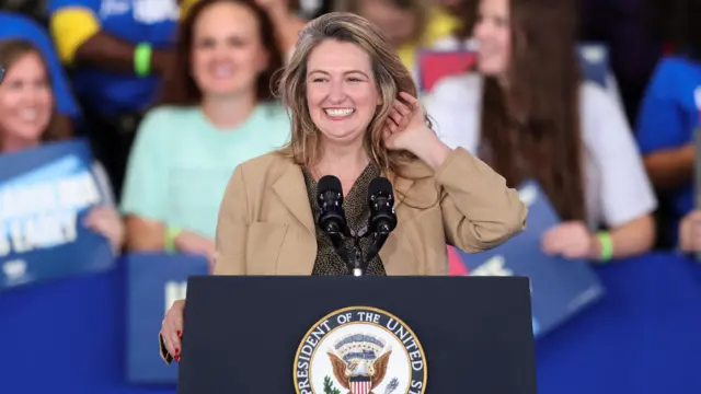 Jennifer Bell smiles as she adjusts her hair at a lectern at the rally with people stood behind her