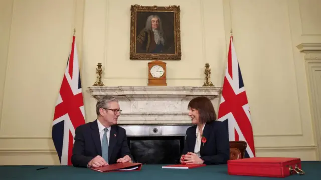 Starmer and Reeves sitting together, with a ministerial red box in front of them, and flags behind