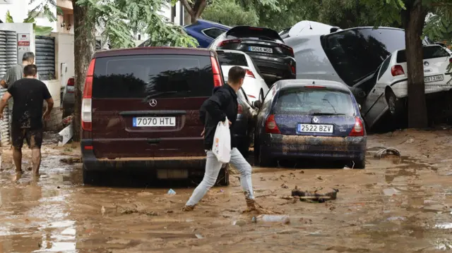 A man walks through a muddy watery road and looks to the right where a stack of cars sit