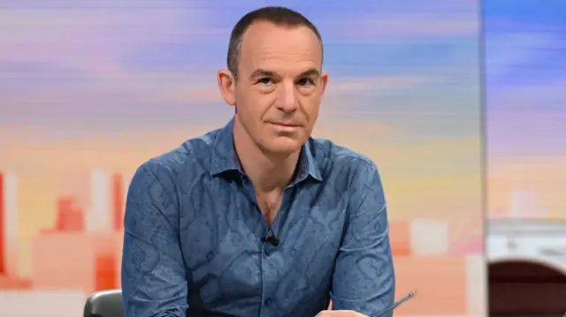 Martin Lewis sat at a desk with his arms resting on it. He holds a pen in one hand. There is a glass of water in front of him