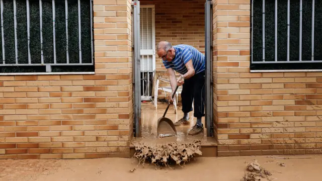 A man uses a dustpan to remove muddy water from the front of his property