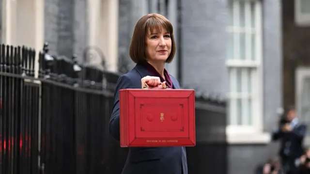 Rachel Reeves outside Number 10 ahead of Budget. She's holding a red ministerial box containing the new measures. Reeves is wearing a dark blue suit with burgundy blouse