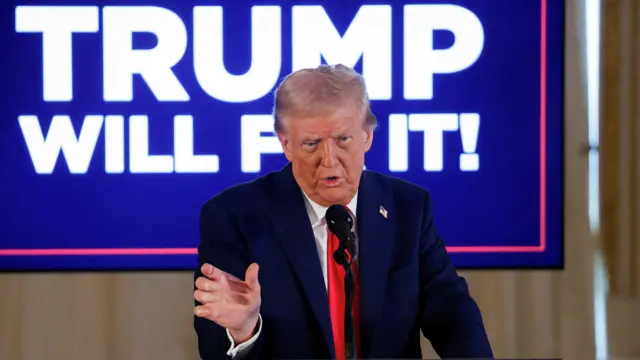 Donald Trump stands in front of a blue 'Trump will fix it!' sign speaking into a microphone