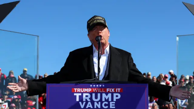 Donald Trump stands on a stage in front of a podium while holding his arms out. A crowd is behind him. He is wearing a black baseball cap with the words Make America Great Again written on it. He is speaking into a microphone.