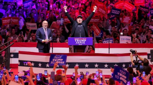 Elon Musk on stage with both hands raised in the air as he appears to shout. He is surrounded by a sea of people holding Trump signs and phones up in the air