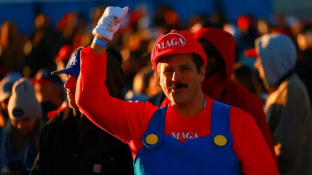 A man wearing a Mario costume and a MAGA shirt underneath with a thumbs up in the air