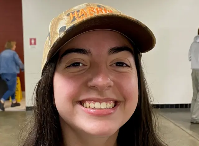 A young woman with dark hair in a camaflogue pattern Harris-Walz hat