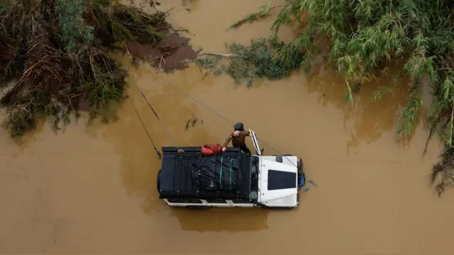 A partially submerged SUV with bags strapped to the roof is seen in brown water. A man is opening the driver's side door. Downed trees frame the truck.