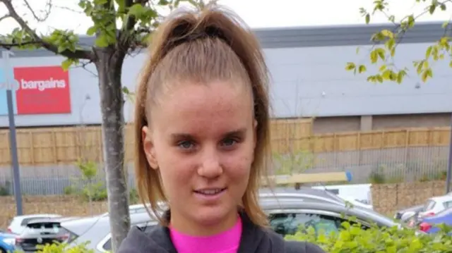 Holly Newton standing in a car park. She has long dark hair and is wearing a pink t shirt