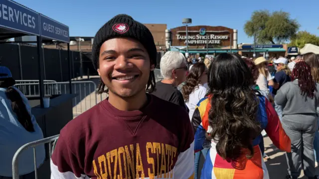 Young Black voter poses smiling for photo. He's wearing a black woollen hat, an Arisona State sweatshirt in burgundy, white and yellow. Behind him is a group of people queuing outside the Talking Stick Resort Amphitheatre