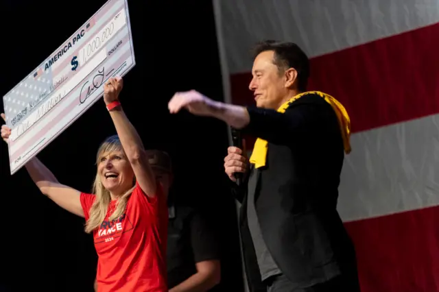 Elon Musk in dark suit with yellow cloth over shoulder next to woman in red shirt holding up oversized check