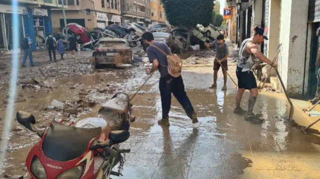 People cleaning up mud off the street, with a pile of cars behind them