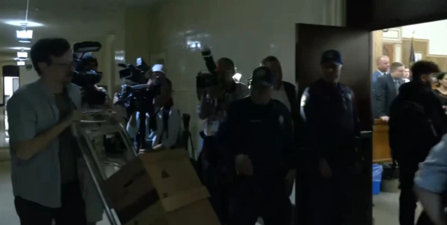 Cameramen outside court as a person walks past with boxes