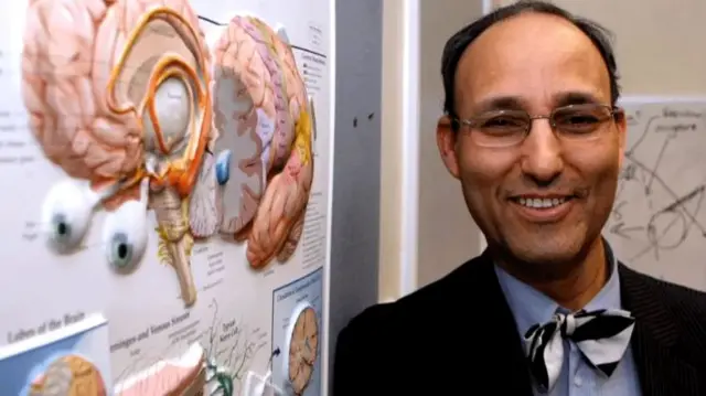 Prof Sam Eljamel - smiling man in a suit and bowtie, standing next to a wall showing diagrams of the brain