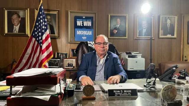 Kevin Moore sits at a desk with an American flag and a Harris poster in the background