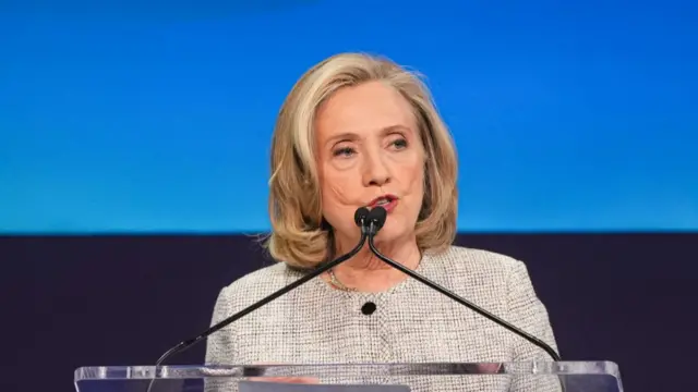 Hillary Clinton speaks on stage in white and black suit with black buttons. Blue background