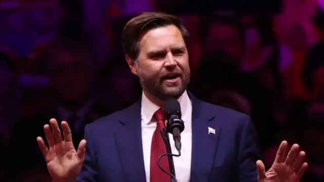 JD Vance speaks at rally wearing dark blue suit with white shirt and deep red tie, US flag pin on left lapel. Both hands are raised and blurred background of crowd