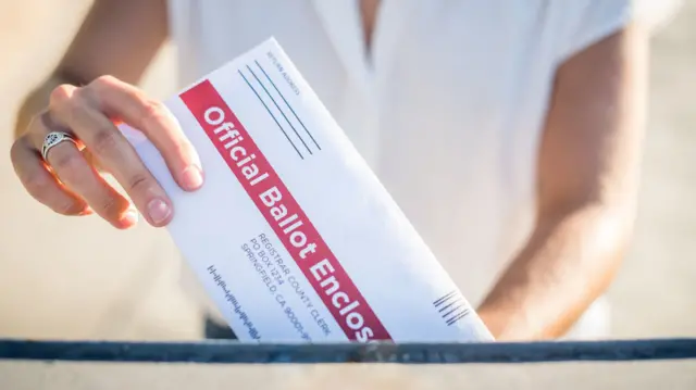 Postal vote being cast through post box. Woman's hand with ring on right ring finger, blurred background is a blurred woman in white short sleeved shirt whose head is out of shot