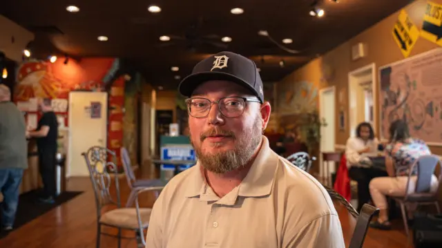 William Greene is sitting in a cafe. He wears a black cap with the letter D in Gothic font on the front, a beige polo shirt and glasses.