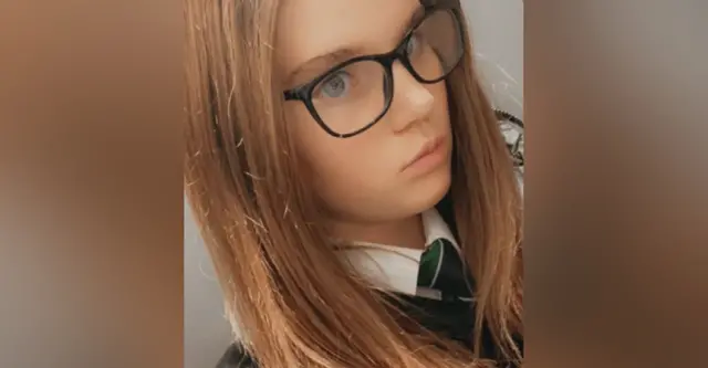 Holly Newton takes a selfie, she is wearing a school uniform with a white shirt and green and blue striped tie, black glasses and has long light brown hair