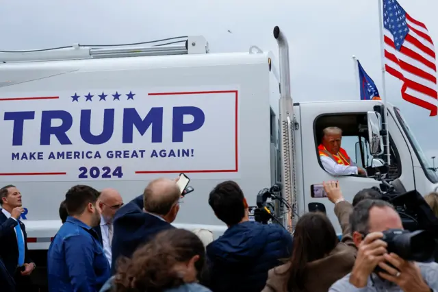 Trump leans out a Trump-branded garbage truck with reporters