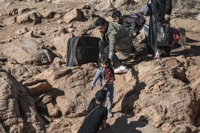 Adults and children climbing over rocks carrying bags and suitcases