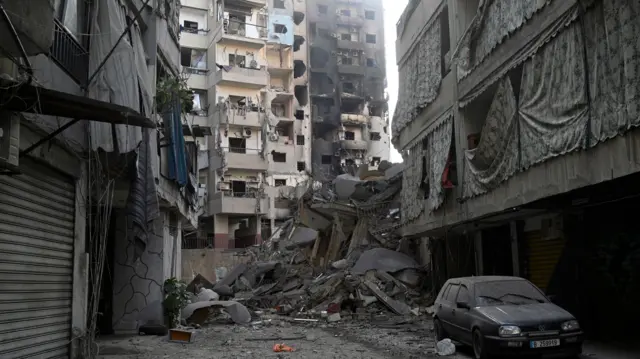 A view of a street with a collapsed building at the end and burnt out buildings still standing in the background