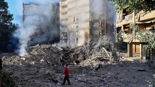 A man walks among rubble with damaged buildings in the background and smoke rising from the ground