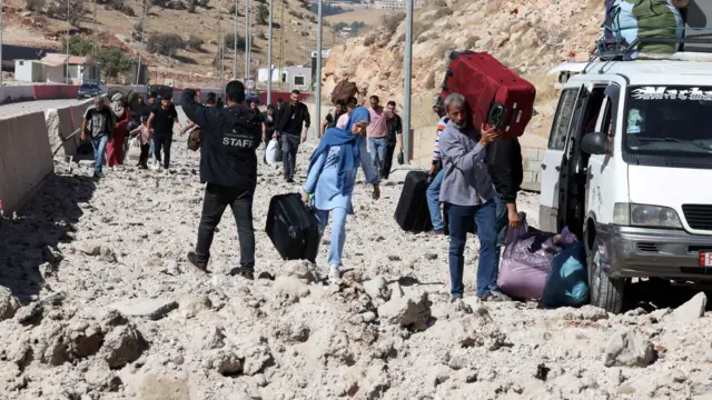 People walk along a road full of rubble carrying suitcases and bags