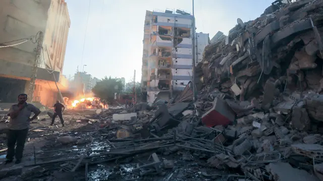 Rubble strewn streets, a flat block partly bombed and exposed, and people mill about in the street in front of a large fire