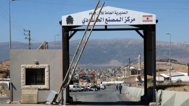 A border crossing between Syria and Lebanon. A black arch frame has Arabic writing across the top. People and cars can be seen in the background.