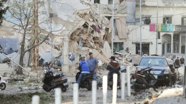 Men stand around with motorbikes and cars - a collapsed building can be seen in the background