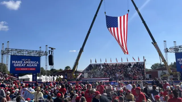 A crowd at Trump's rally looks at the stage, surrounded by bulletproof glass, with an American flag flying overhead