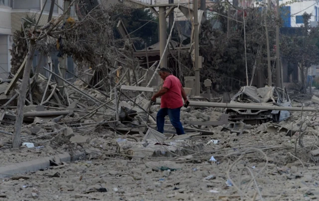 A man walks on the rubble of a damaged site, in the aftermath of Israeli strikes on Beirut's southern suburbs. Debris can be seen including broken pieces of metal and rubble. A car is damaged in the far distance too