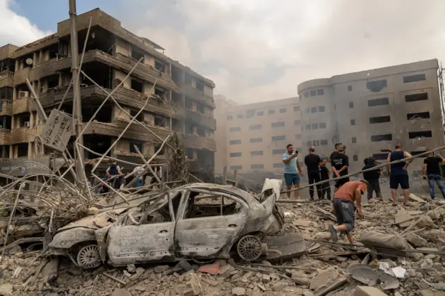 The site of an air strike is seen, with people standing around a crater and burnt out cars