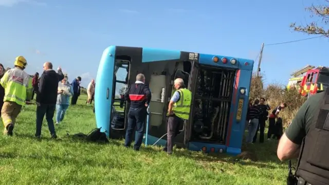 A bus on its side in a field surrounded by school pupils