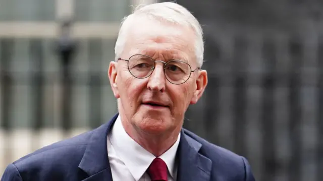 Hilary Benn outside wearing a blue suit and red tie. He is wearing glasses.