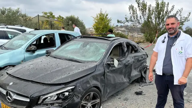 A man in a white doctors jacket stood next to a black Mercedes which has been damaged in an airstrike. There is a green car behind, which has been slightly damaged